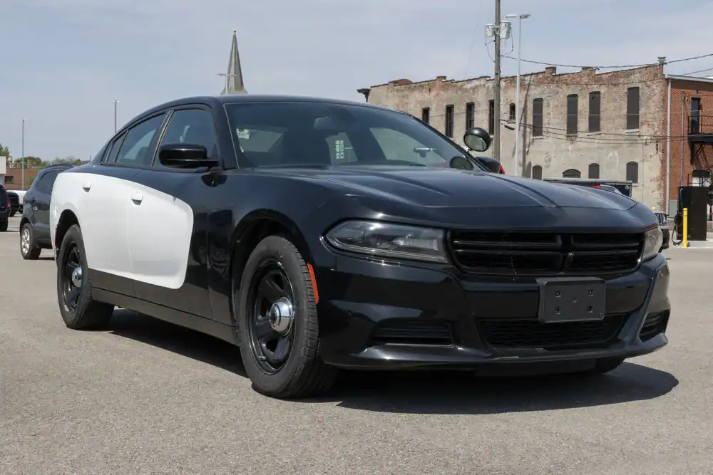 A black and white police car, likely a Dodge Charger, is parked on an empty street with a brick building in the background under a cloudy sky, typical of used police cars NJ or police interceptors NJ found across NY and PA.