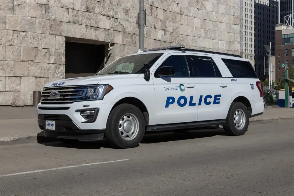 A white Cincinnati Police SUV, similar to used police cars NJ agencies seek, is parked on a city street beside a stone building, with tall buildings rising in the background.