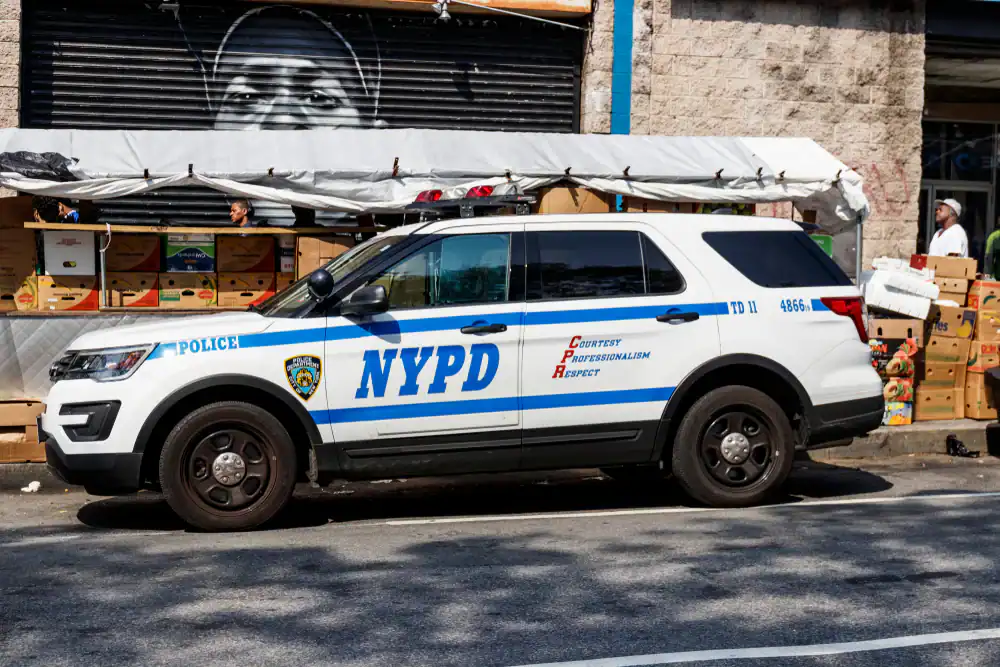 A white NYPD police SUV, similar to police interceptors NJ and used police cars in NJ, NY and PA, is parked on the street beside fruit stands, with a mural of a face visible on a wall in the background.