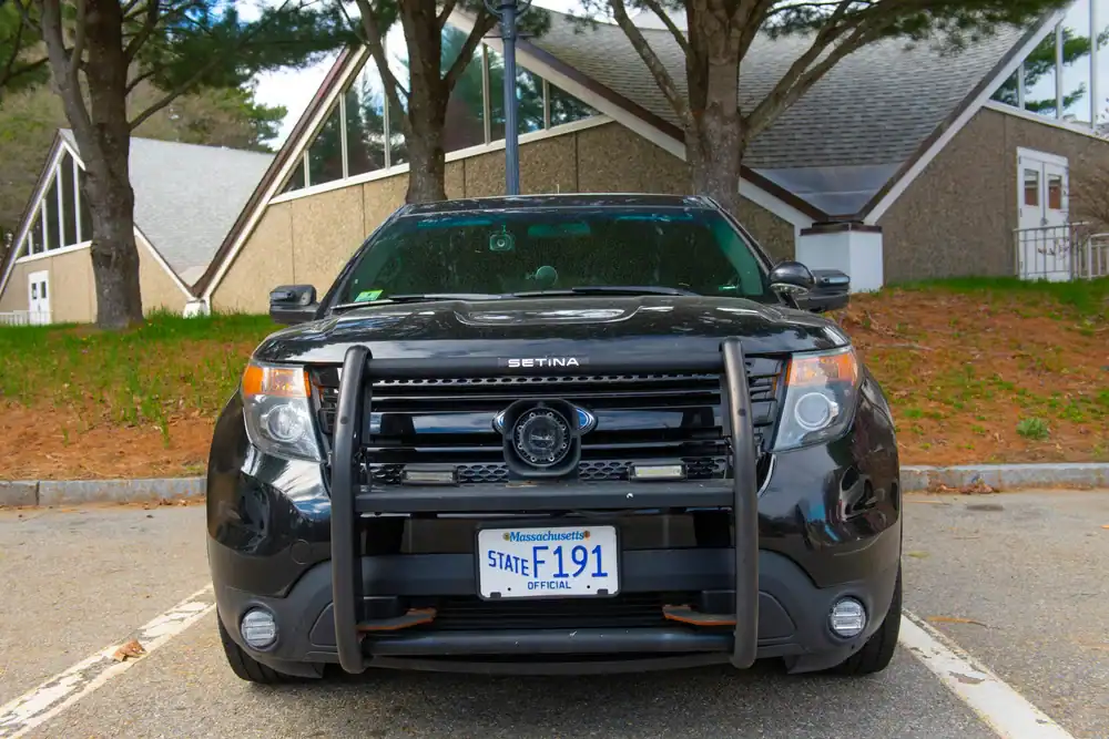 A black police SUV with a bull bar, often found among used police cars NJ dealers offer, is parked in front of a building with large windows. The Massachusetts official state license plate reads "F191." Pine trees and a lamp post are in the background.