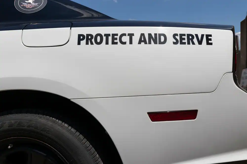 The side of a white police car, often seen among used police cars NJ, with "PROTECT AND SERVE" in bold black letters above the rear wheel, partially showing a tire and a red reflector.
