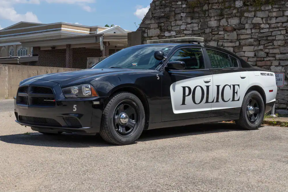 A black and white police car, similar to used police cars NJ agencies drive, is parked on a paved surface near a stone wall and a building with columns. The word "POLICE" is printed in large letters on the car's side.