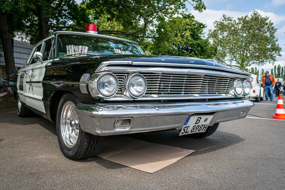 A vintage black and white Ford police car, a classic example of police interceptors NJ,NY and PA, is parked outdoors on pavement. Its front view is visible with a red siren atop, trees and people filling the background.