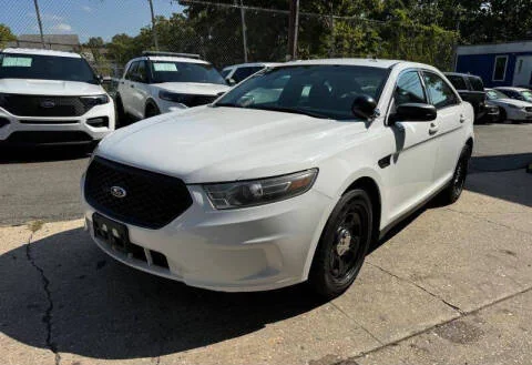 A white Ford sedan, similar to used police cars NJ and police interceptors NJ, sits on a paved area beside a chain-link fence, with other vehicles in the background on a sunny day.