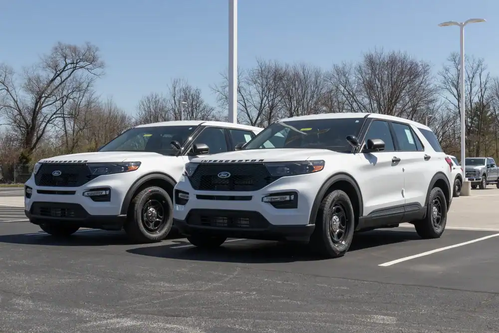 Two white Ford Explorer SUVs, likely former police interceptors NJ, are parked side by side in a lot on a clear day, with bare trees and light poles in the background.